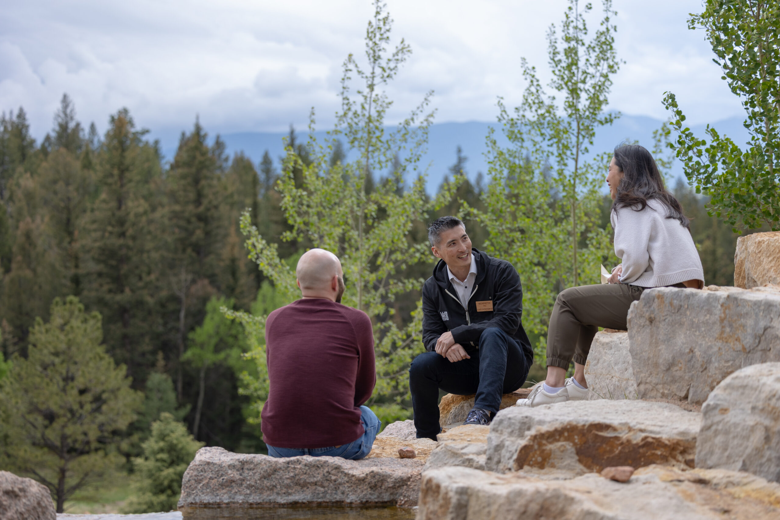 Three adults sit on large stone steps outdoors, talking together against a backdrop of trees and distant mountains on a cloudy day.