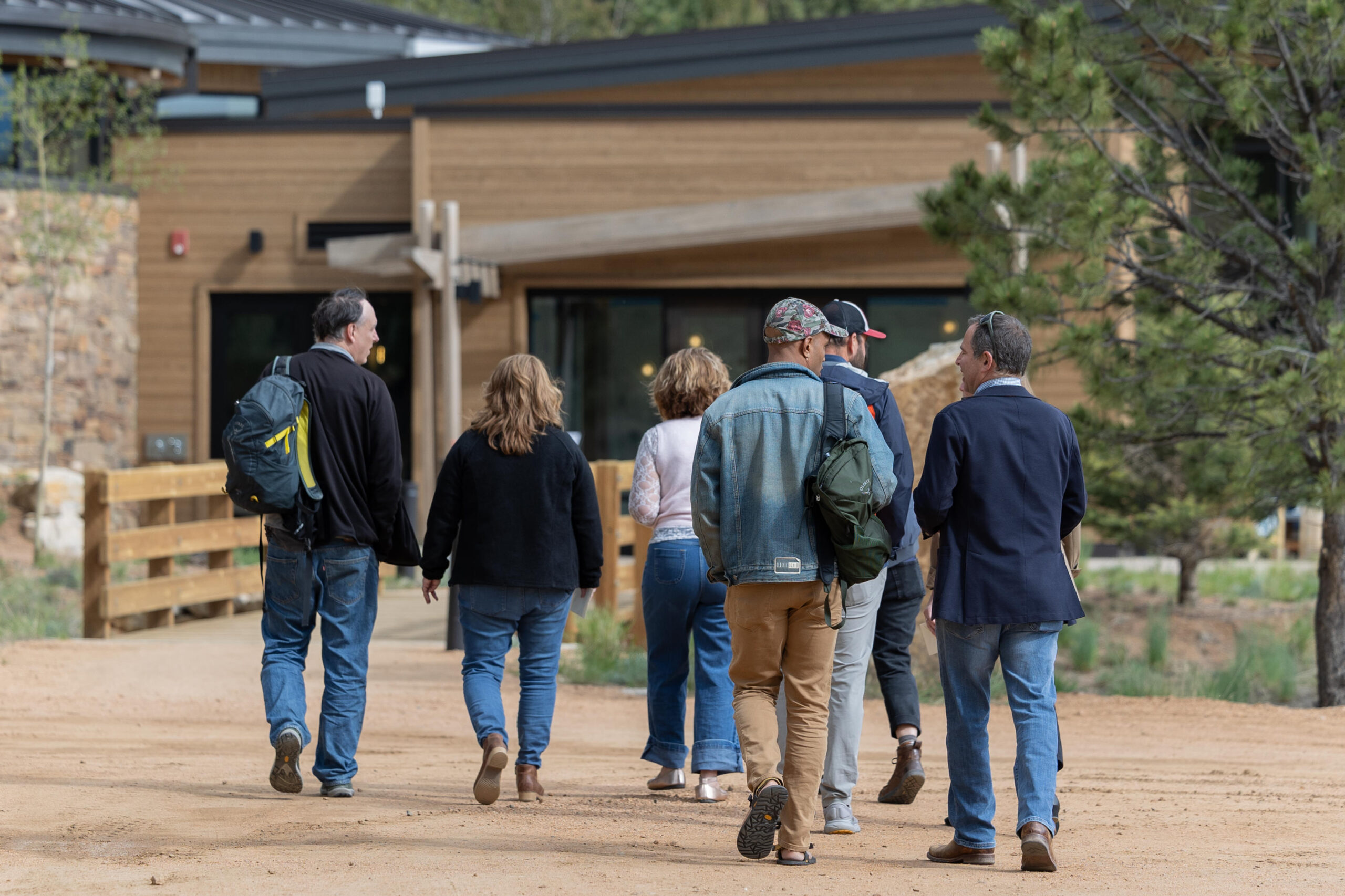 A group of adults walks together along a dirt path toward a modern lodge-style building, carrying backpacks and jackets on a mild day.
