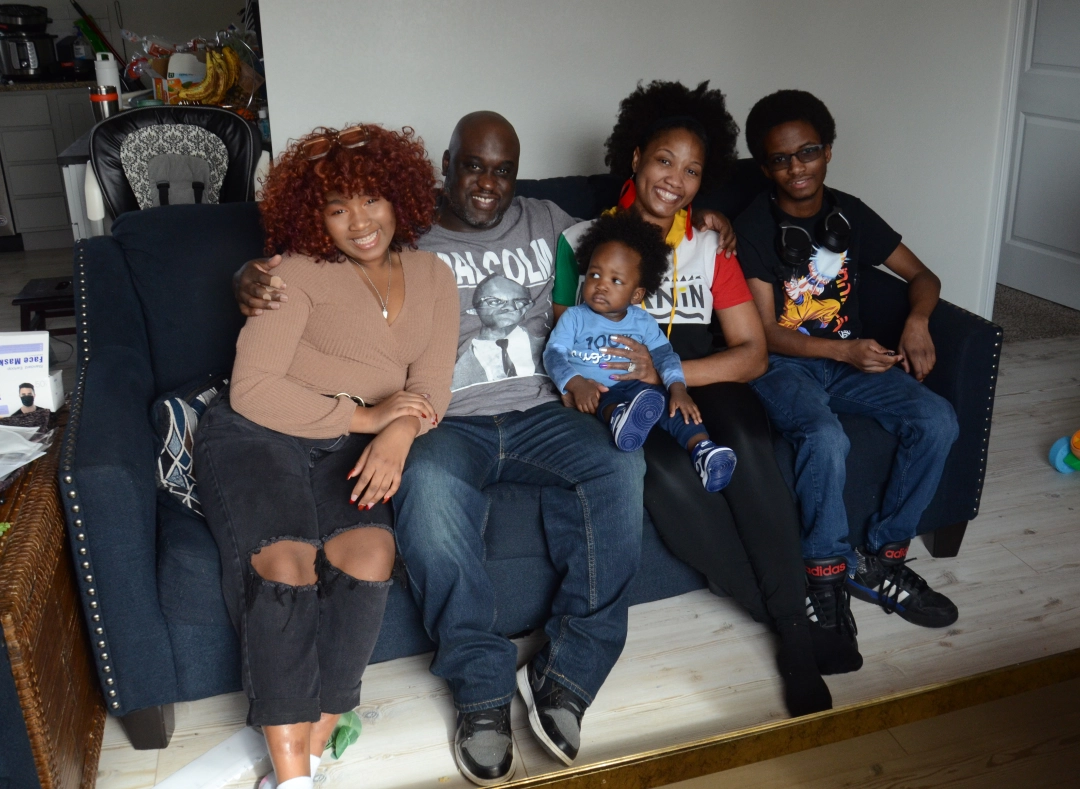 A family of five sits together on a couch, smiling at the camera. Two adults sit in the middle with a toddler on one lap, and two older children sit on either side, creating a warm, relaxed family portrait in their living room.