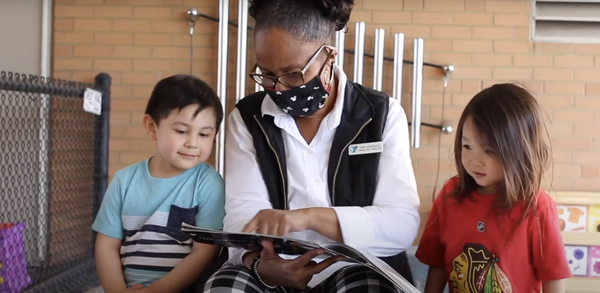 An early childhood teacher wears a mask while reading to two young children