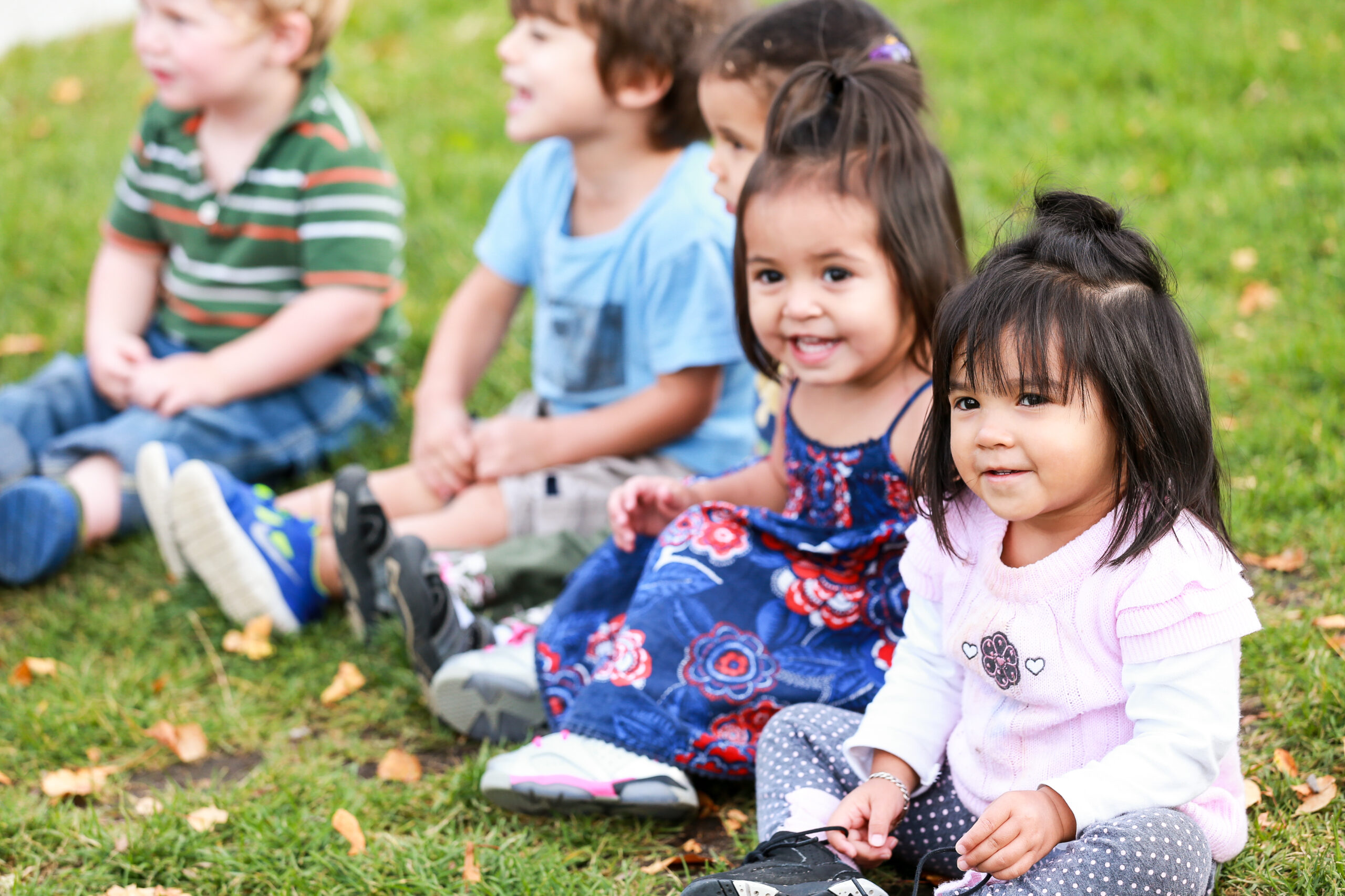 Young children sit together on the grass outdoors, smiling and watching something off-camera. The scene is bright and playful, with kids dressed in colorful outfits.