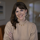 A woman smiles at the camera in an indoor setting, wearing a light sweater and statement earrings.