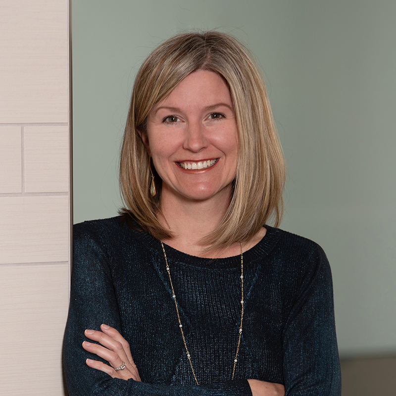 A woman with blonde shoulder-length hair smiles at the camera with her arms crossed, standing against a light-colored wall.