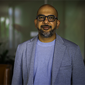 A man wearing glasses, a light blazer, and a blue shirt stands indoors, looking at the camera with a neutral expression.
