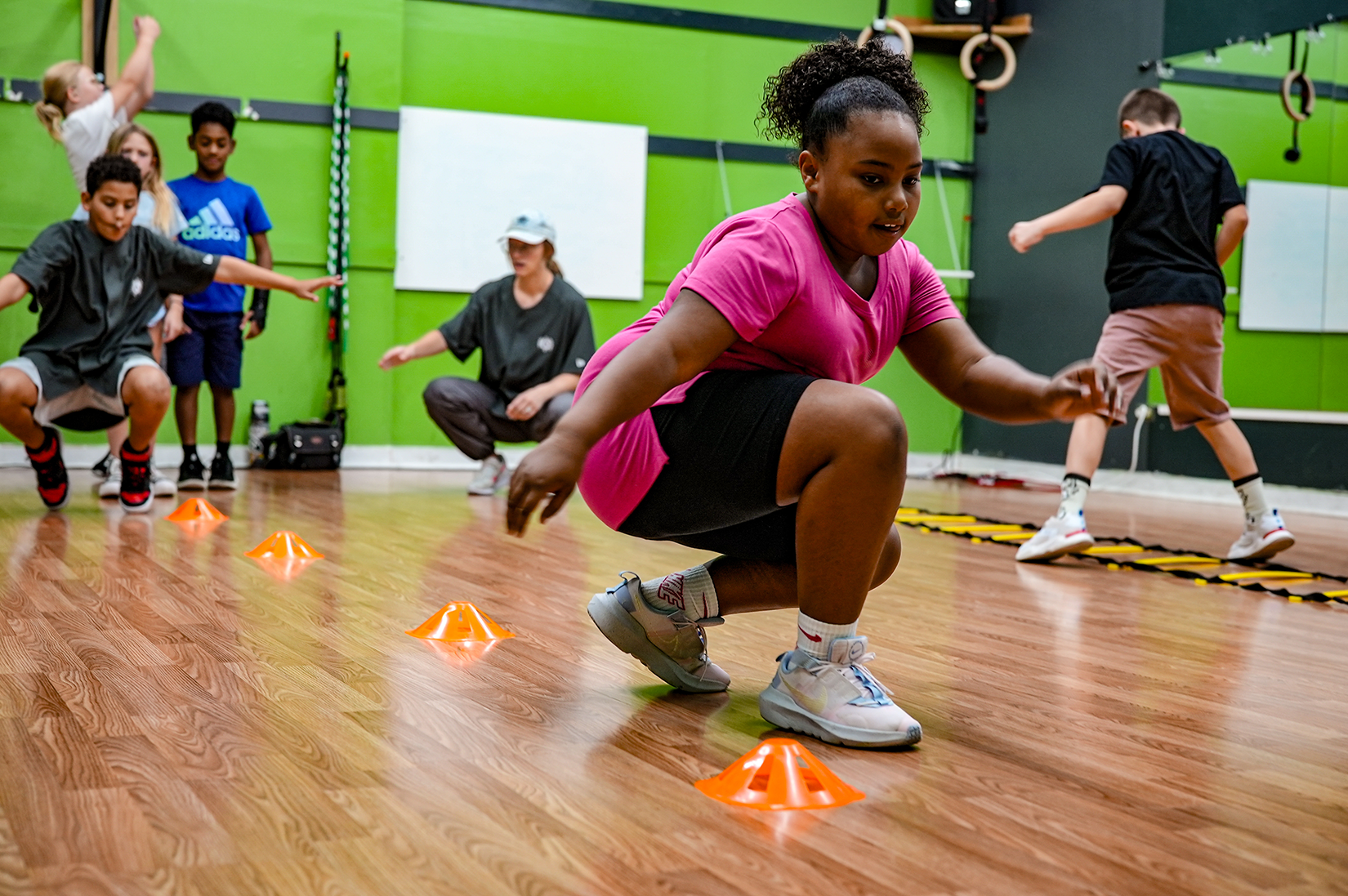 A group of kids participates in break dancing exercises at the School of Breaking, with one girl in a pink shirt leading as she moves between orange cones on the gym floor.