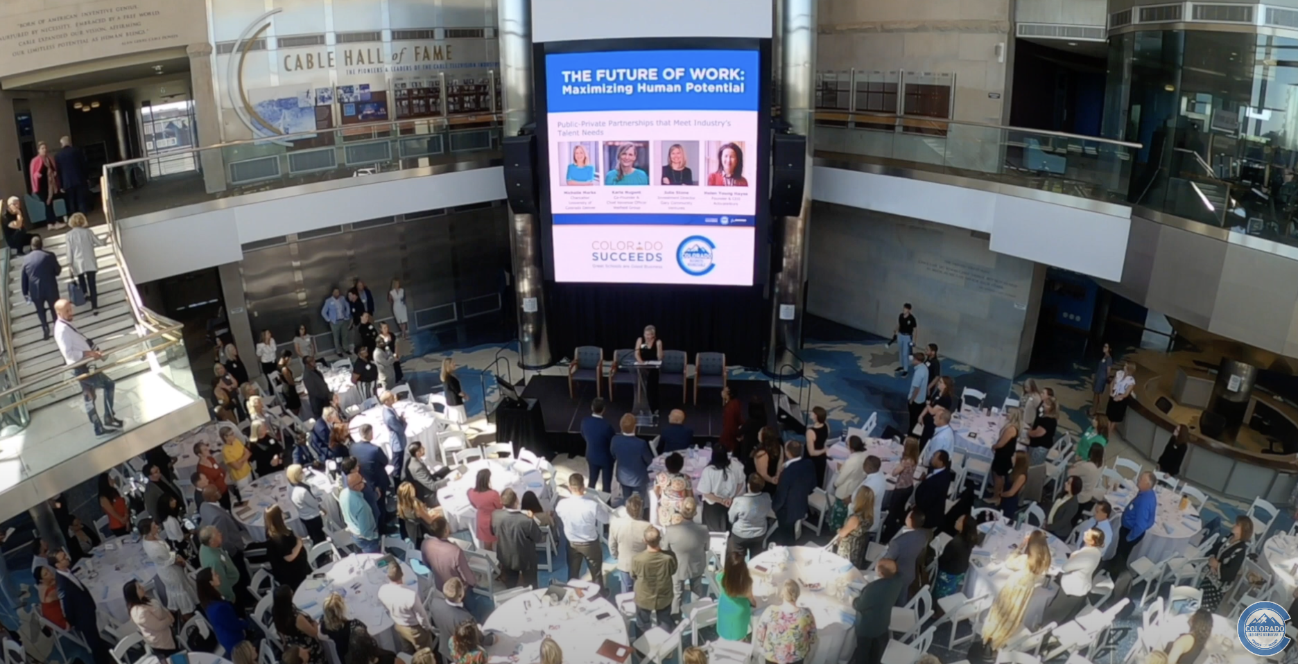 A large crowd gathers around round tables in a spacious event hall as a speaker stands at a podium on stage. A giant screen above the stage displays “The Future of Work: Maximizing Human Potential” along with panelist photos and event branding.