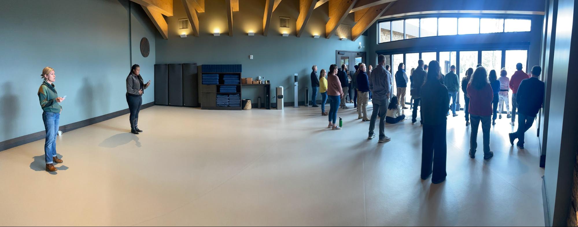 A group of people stands near large windows listening to two presenters at the front of a spacious room with high ceilings and natural light.