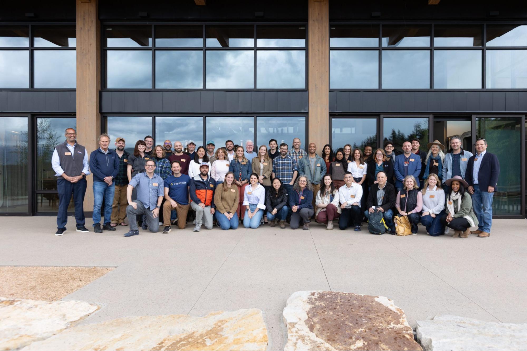 A large group of adults poses for a photo outside a modern building with tall windows. They stand and kneel in several rows, smiling toward the camera, taken during the Activate40 retreat.